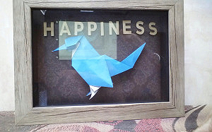 Origami bird sits in a small shadow box. small letters above it spell out happiness